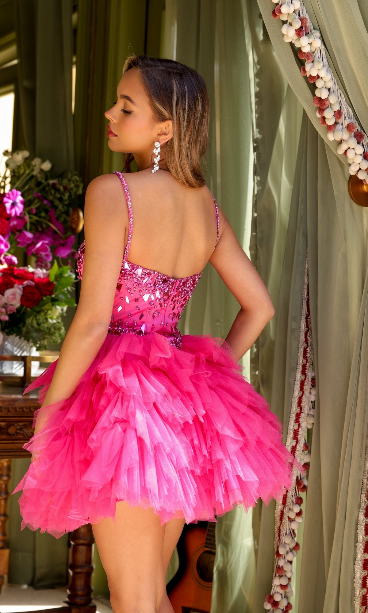 A woman stands with her back turned, wearing the Ava Presley 29180 Short Tiered Homecoming Dress in bright pink with ruffles and sequins. She is indoors, close to green curtains and a bouquet of flowers.